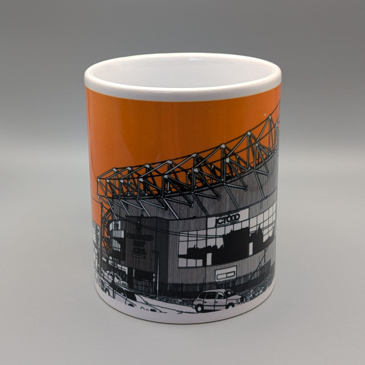 A white ceramic mug featuring a printed graphic of the Old Trafford stadium, with a solid orange background on the upper half of the design and a black-and-white architectural illustration below.