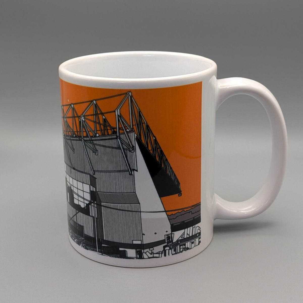 A white ceramic mug featuring a graphic print of a stadium structure in black and gray against a solid orange background, set against a neutral gray studio backdrop.