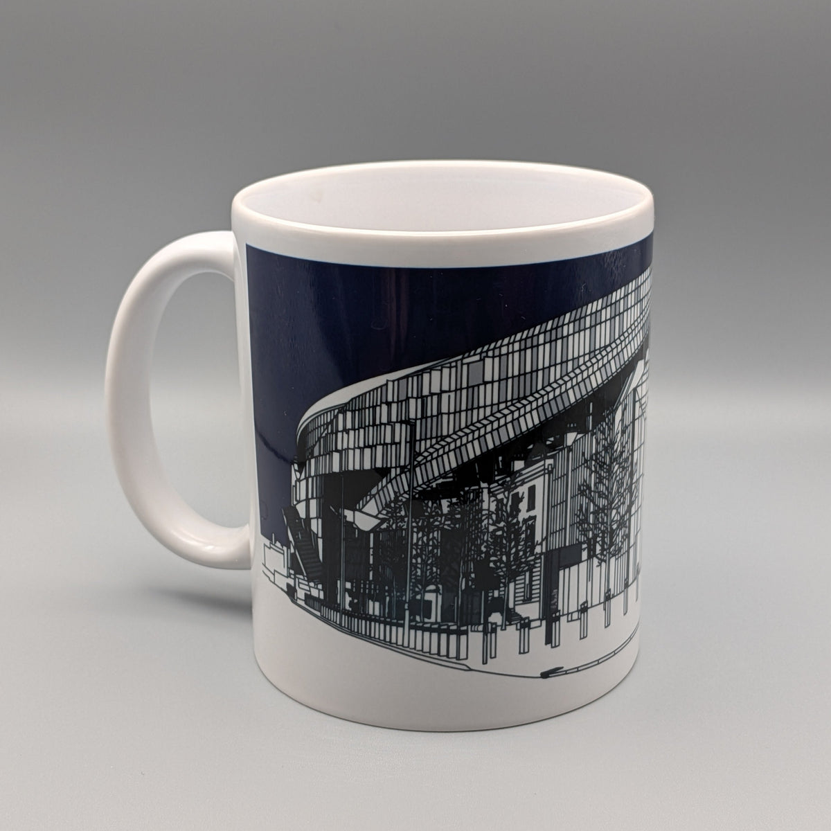 A white ceramic mug featuring a dark navy blue wrap-around graphic print of a modern architectural stadium or arena, shown on a neutral gray studio background.