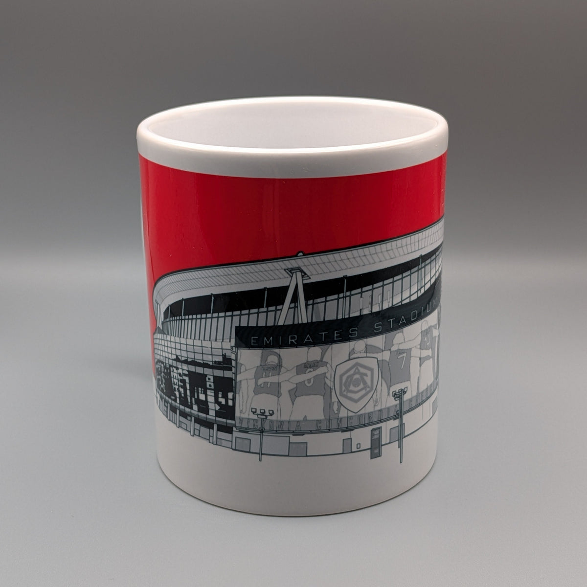 A white ceramic mug featuring a bold red band and a black-and-white graphic illustration of Emirates Stadium, set against a neutral gray studio background.