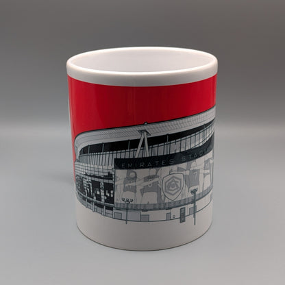 A white ceramic mug featuring a bold red band and a black-and-white graphic illustration of Emirates Stadium, set against a neutral gray studio background.