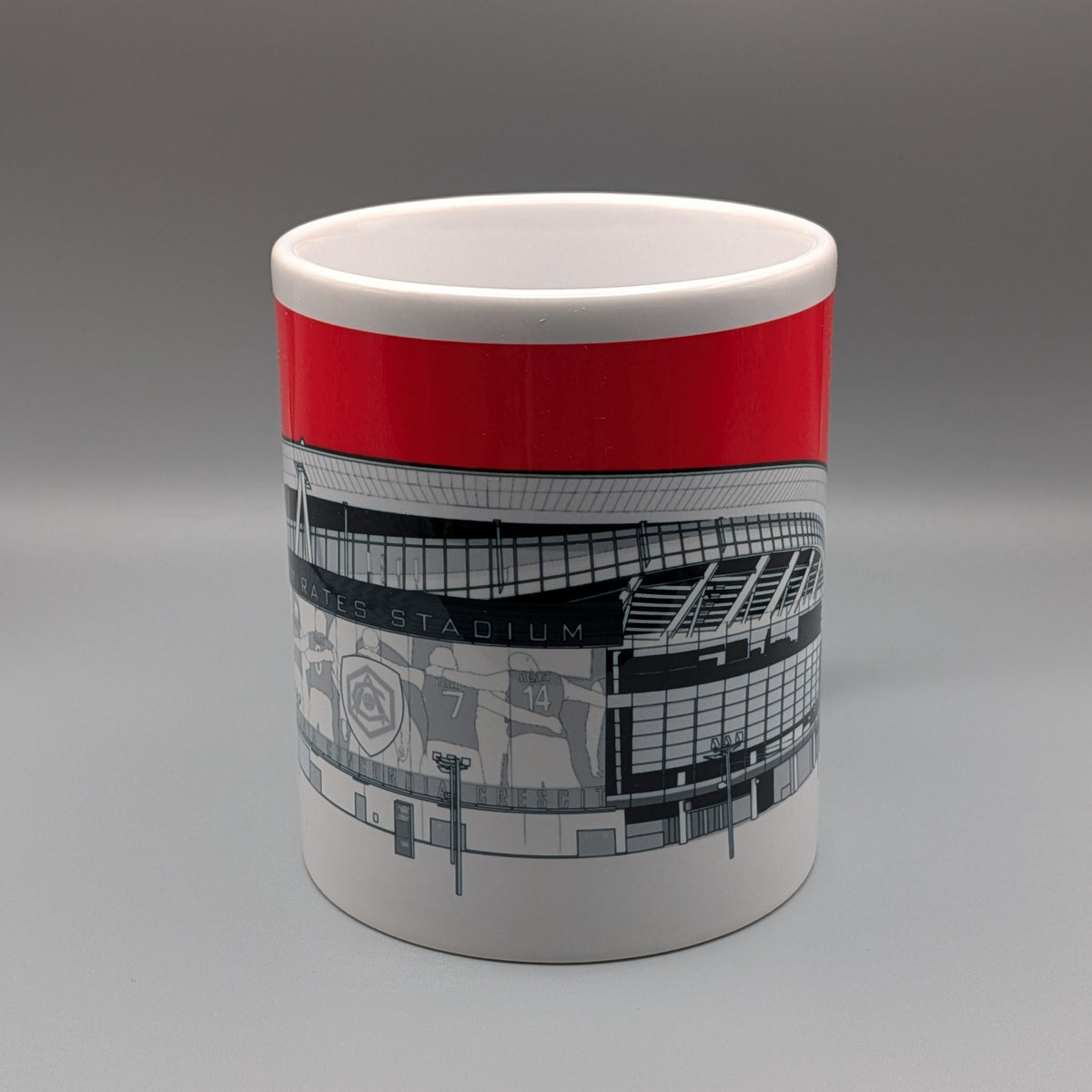 A white ceramic mug featuring a bold red band around the upper half and a black-and-white architectural illustration of a stadium on the lower half, shown against a neutral gray studio background.