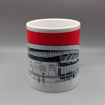 A white ceramic mug featuring a bold red band around the upper half and a black-and-white architectural illustration of a stadium on the lower half, shown against a neutral gray studio background.