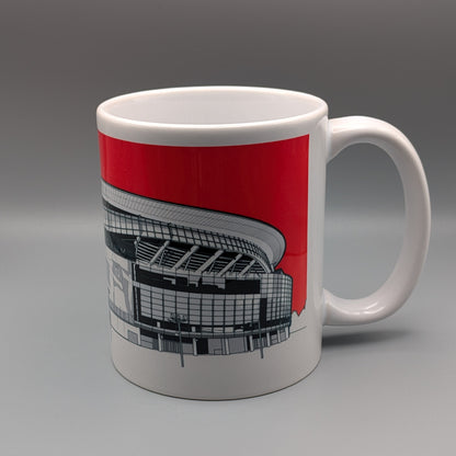 A white ceramic mug featuring a graphic print of a stadium under a bold red sky, set against a neutral gray studio background.